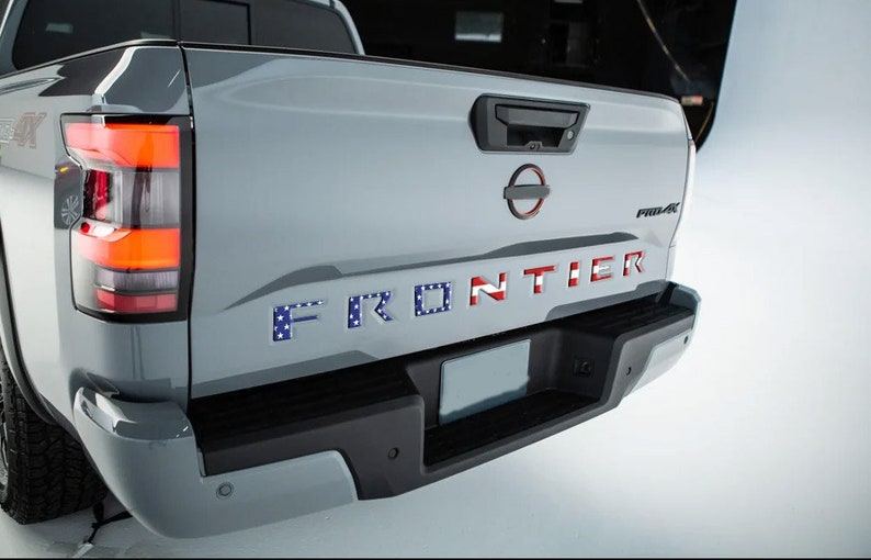 May include: A gray pickup truck with a black bumper and a red, white, and blue "FRONTIER" emblem on the tailgate. The truck has a black tailgate handle and a black Nissan emblem.