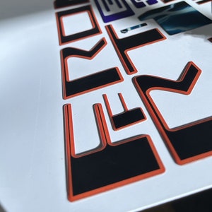 May include: Close-up of black and orange vinyl lettering that spells out "FORD" in a stylized font. The letters are raised and have a glossy finish.
