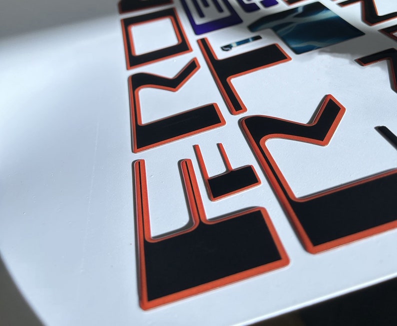May include: Close-up of black and orange vinyl lettering that spells out "FORD" in a stylized font. The letters are raised and have a glossy finish.