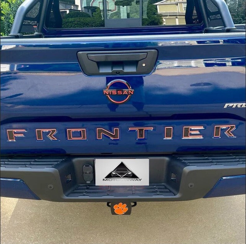 May include: A blue Nissan Frontier pickup truck with a red "FRONTIER" lettering on the tailgate. The truck has a black bumper with a silver "MOTORWAY" license plate and a black tow hitch with an orange paw print.