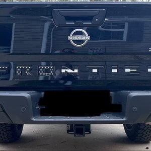 May include: Black Nissan Frontier pickup truck with a black and white American flag design on the tailgate lettering. The truck has a black bumper with a silver hitch receiver.
