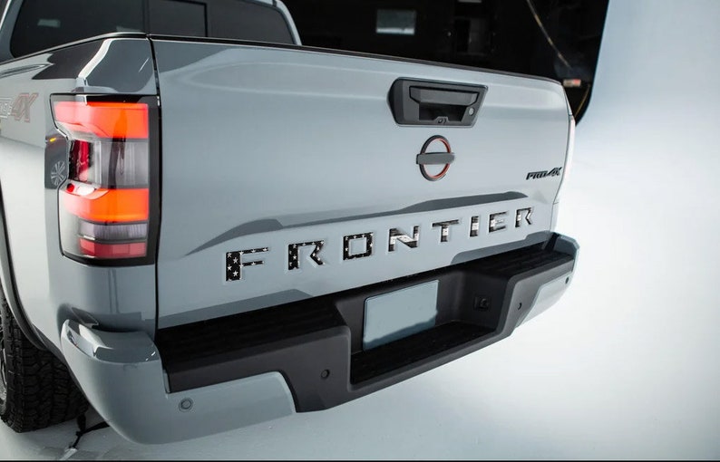 May include: A gray pickup truck with a black tailgate and a black "FRONTIER" emblem with a stars and stripes pattern. The truck has a black bumper with a license plate holder and a black rearview camera.