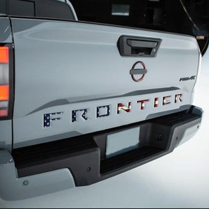 May include: A gray pickup truck with a black bumper and a license plate. The truck has a "FRONTIER" tailgate with an American flag design.