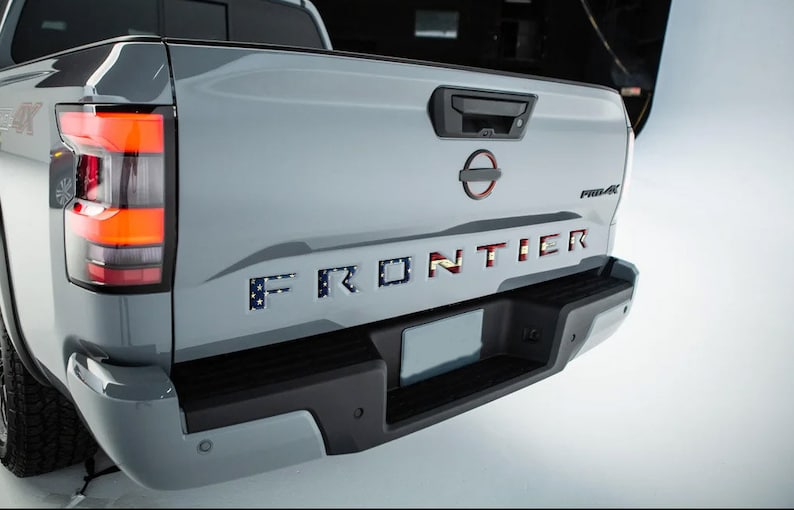 May include: A gray pickup truck with a black bumper and a license plate. The truck has a "FRONTIER" tailgate with an American flag design.