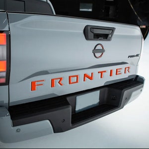 May include: A gray pickup truck with a black bumper and a red "FRONTIER" emblem on the tailgate. The truck has a black tailgate handle and a Nissan logo.