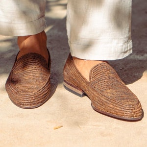 Può includere: Un paio di mocassini marroni in pelle intrecciata con suola marrone chiaro. I mocassini sono indossati da una persona che indossa pantaloni bianchi.