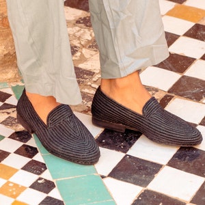 Pode incluir: Um par de sapatos de couro preto tecido com bico fino. Os sapatos são usados por uma pessoa que usa calças azuis claras e estão em um chão de ladrilhos preto e branco.