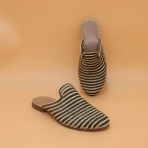 May include: Two woven, open-back slippers with black and beige horizontal stripes. The slippers have a rounded toe and a low heel. One slipper is lying flat, and the other is upright, showing the interior lining.