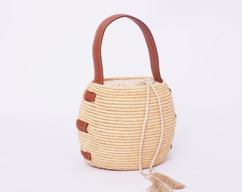 May include: A straw bucket bag with a brown leather handle and tassels. The bag has a woven texture and is a light beige color.