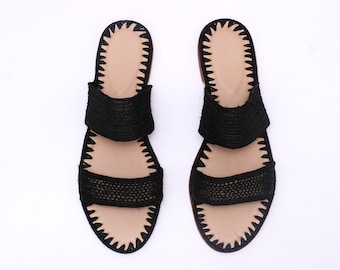Op de afbeelding: Een paar zwarte geweven sandalen met twee banden over de bovenkant. De sandalen hebben een bruine leren zool en een gecrochete rand.