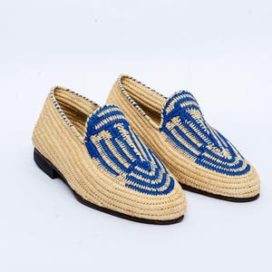 May include: A pair of woven raffia loafers. The shoes have a natural beige color with blue geometric designs on the top. The soles are dark brown. The shoes are displayed on a white background.