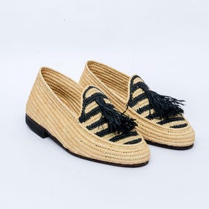 Puede incluir: Un par de mocasines de rafia tejida. Los zapatos son de color beige con detalles de rayas negras en la pala y borlas negras. Las suelas son de color marrón oscuro.