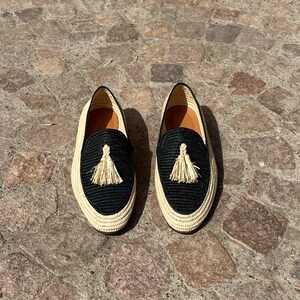 Puede incluir: Un par de mocasines tejidos negros y crema con un detalle de borla. Los mocasines est&aacute;n sobre una superficie de piedra.
