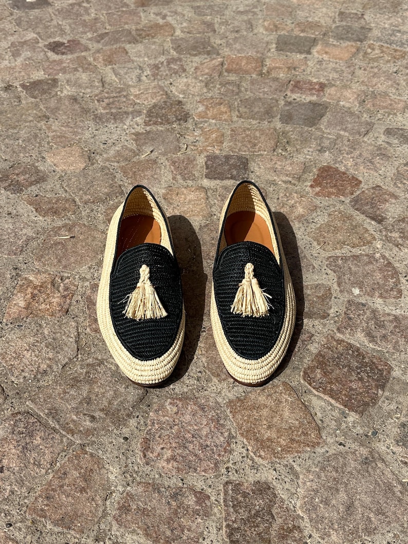 Puede incluir: Un par de mocasines tejidos negros y crema con un detalle de borla. Los mocasines est&aacute;n sobre una superficie de piedra.