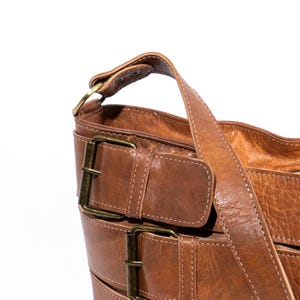 Pu&ograve; includere: Una borsa a tracolla in pelle marrone con pi&ugrave; fibbie e una lunga tracolla.