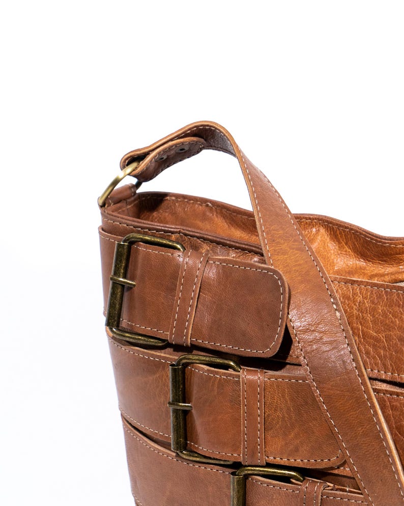 Pu&ograve; includere: Una borsa a tracolla in pelle marrone con pi&ugrave; fibbie e una lunga tracolla.