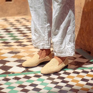 May include: A pair of beige woven loafers with a rounded toe. The shoes are worn with white linen pants. The floor is tiled with a pattern of black, white, yellow, and green squares.