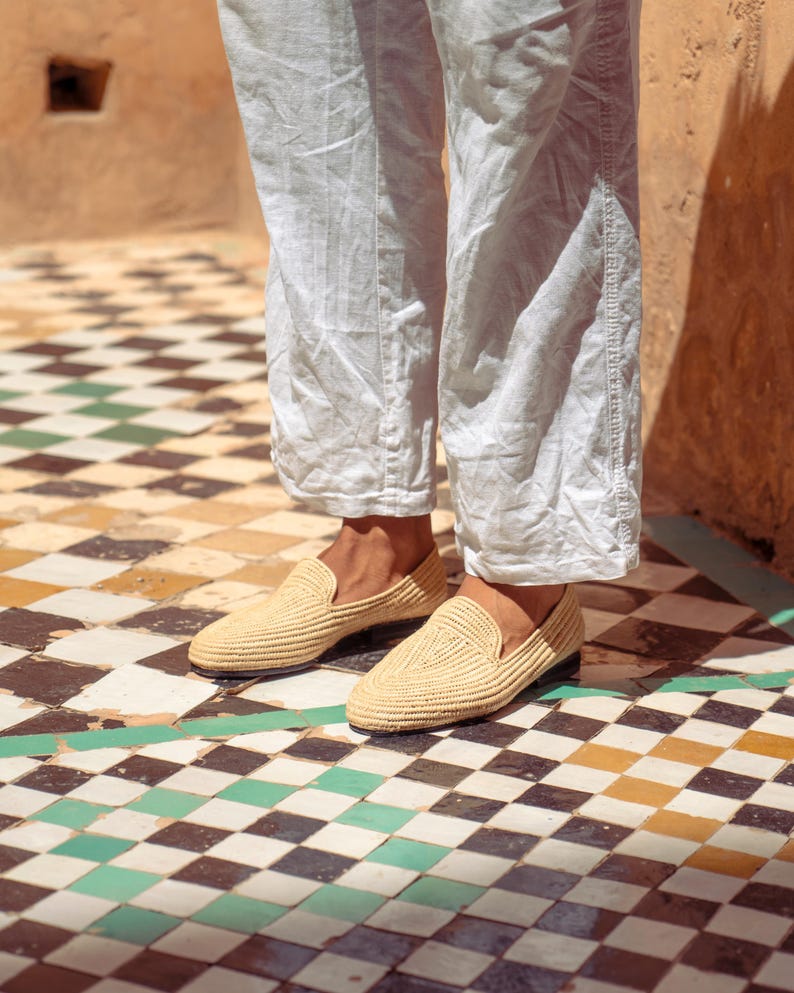 May include: A pair of beige woven loafers with a rounded toe. The shoes are worn with white linen pants. The floor is tiled with a pattern of black, white, yellow, and green squares.