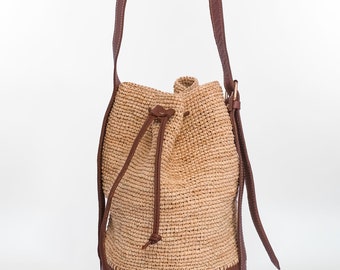 May include: A beige woven bucket bag with a brown leather base and drawstring closure. The bag has a brown leather strap and a gold-colored metal buckle. The bag is approximately 10 inches tall.