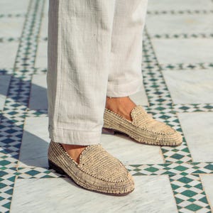 Pode incluir: Um par de mocassins de palha tran&ccedil;ada de cor castanho claro. Os mocassins s&atilde;o usados por uma pessoa que usa cal&ccedil;as brancas.