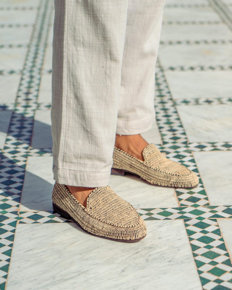 Pode incluir: Um par de mocassins de palha tran&ccedil;ada de cor castanho claro. Os mocassins s&atilde;o usados por uma pessoa que usa cal&ccedil;as brancas.