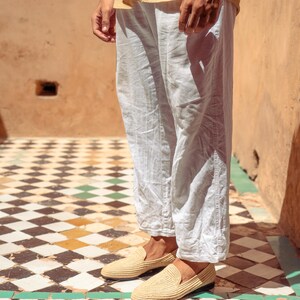 May include: A pair of beige woven loafers. The shoes are worn with light-colored linen pants. The background features a checkered tile floor with black, white, and green tiles, and a peach-colored wall.