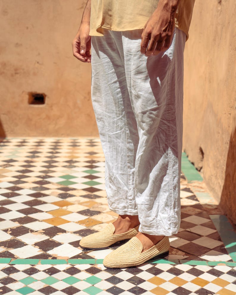 May include: A pair of beige woven loafers. The shoes are worn with light-colored linen pants. The background features a checkered tile floor with black, white, and green tiles, and a peach-colored wall.