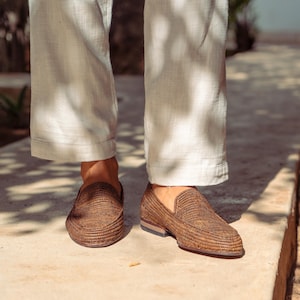 K&ouml;nnte beinhalten: Ein Paar braune geflochtene Lederschuhe mit dezenter Textur. Die Schuhe werden von einer Person getragen, die eine wei&szlig;e Hose tr&auml;gt.