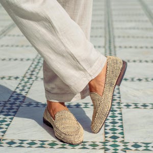 Pode incluir: Um par de mocassins de palha tran&ccedil;ada com detalhes pretos. Os mocassins est&atilde;o sendo usados por uma pessoa em p&eacute; em um piso de ladrilhos.