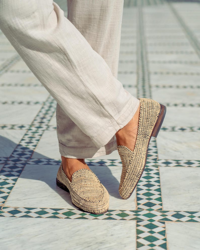 Pode incluir: Um par de mocassins de palha tran&ccedil;ada com detalhes pretos. Os mocassins est&atilde;o sendo usados por uma pessoa em p&eacute; em um piso de ladrilhos.