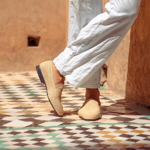 May include: Tan woven loafers with a black sole. The shoes are made of a natural fiber material and are worn with light blue linen pants. The image is taken outdoors on a tiled floor.