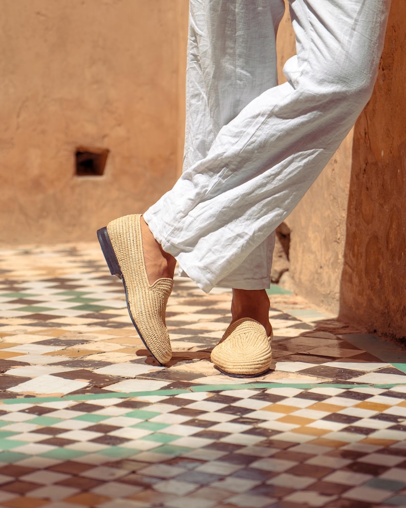 May include: Tan woven loafers with a black sole. The shoes are made of a natural fiber material and are worn with light blue linen pants. The image is taken outdoors on a tiled floor.