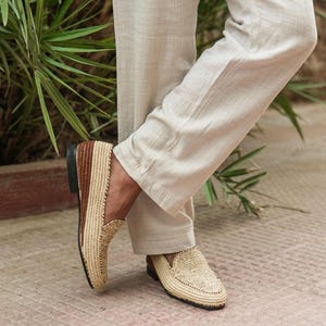 Puede incluir: Un par de mocasines de paja trenzada con detalles de cuero marr&oacute;n. Los mocasines los lleva puestos una persona de pie sobre un suelo de baldosas.