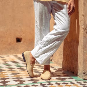 May include: Tan woven loafers with a black sole. The shoes are worn with light-colored linen pants and a yellow shirt. The setting is outdoors on a tiled floor with a geometric pattern.