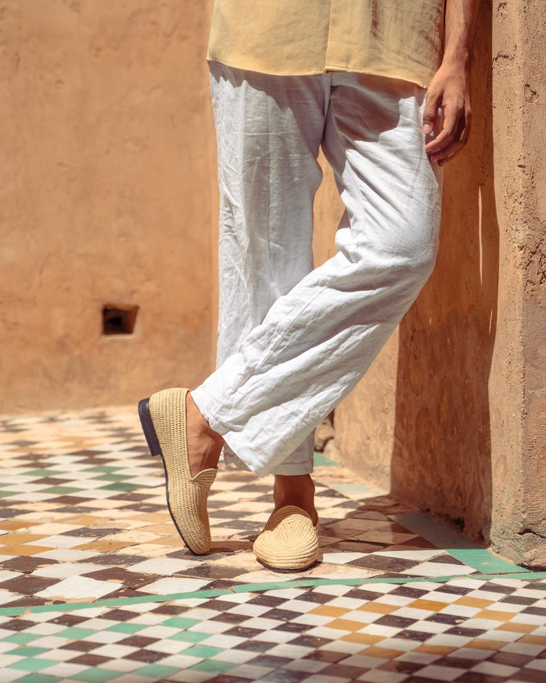 May include: Tan woven loafers with a black sole. The shoes are worn with light-colored linen pants and a yellow shirt. The setting is outdoors on a tiled floor with a geometric pattern.