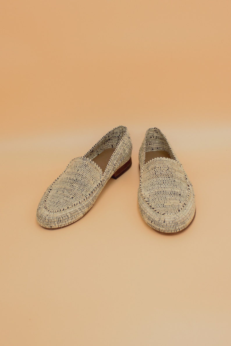 Pode incluir: Um par de mocassins de palha tran&ccedil;ada com acabamento preto. Os mocassins est&atilde;o sobre um fundo bege claro.