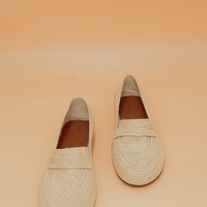 May include: A pair of cream-colored woven straw loafers with brown leather insoles. The loafers are on a light orange background.