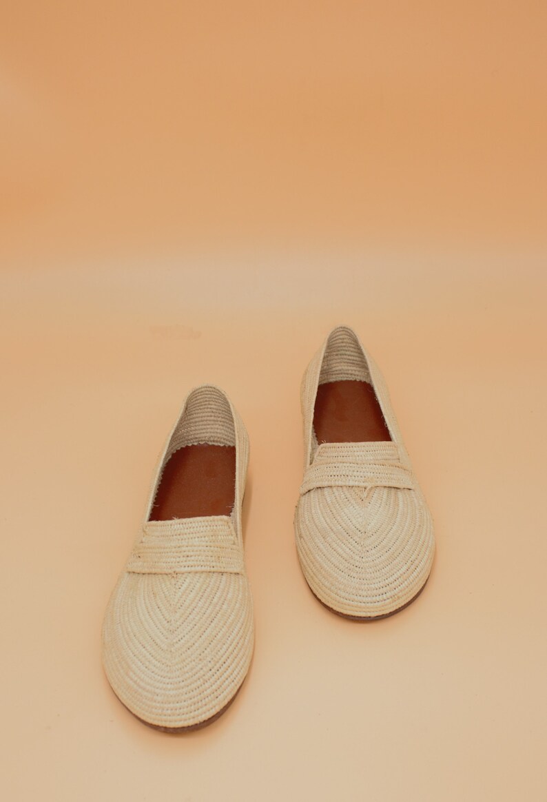 May include: A pair of cream-colored woven straw loafers with brown leather insoles. The loafers are on a light orange background.
