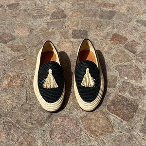 Puede incluir: Un par de mocasines tejidos negros y beige con borlas. Los mocasines tienen una suela plana y una parte superior tejida.