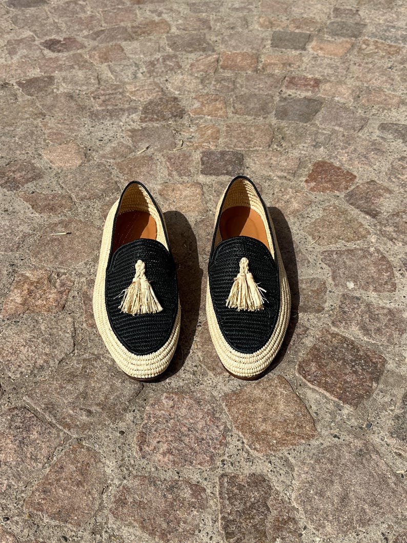 Puede incluir: Un par de mocasines tejidos negros y beige con borlas. Los mocasines tienen una suela plana y una parte superior tejida.