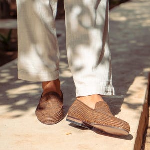 K&ouml;nnte beinhalten: Ein Paar braune geflochtene Lederschuhe mit leicht spitzer Zehenpartie. Die Schuhe werden von einer Person getragen, die eine wei&szlig;e Hose tr&auml;gt.