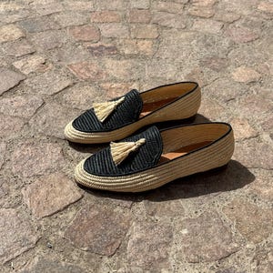 Puede incluir: Un par de mocasines de borlas tejidos en negro y beige. Los mocasines tienen una parte superior tejida con un ribete negro y un detalle de borla en la parte delantera.