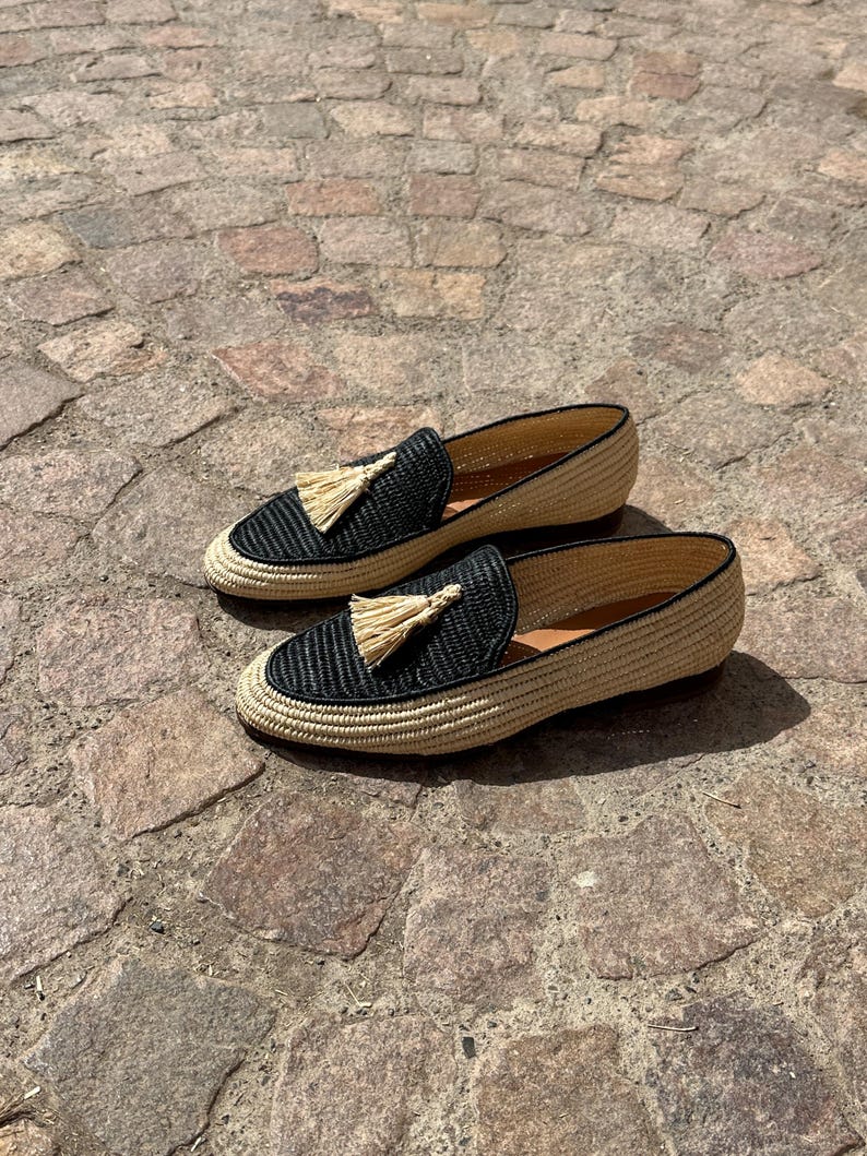 Puede incluir: Un par de mocasines de borlas tejidos en negro y beige. Los mocasines tienen una parte superior tejida con un ribete negro y un detalle de borla en la parte delantera.