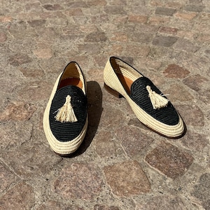 Puede incluir: Un par de mocasines de borlas tejidos en negro y beige. Los mocasines tienen una parte superior tejida en negro con un ribete tejido en beige y una borla beige.