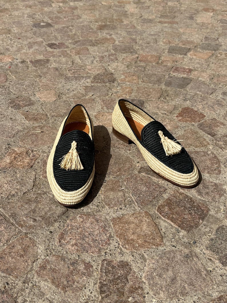 Puede incluir: Un par de mocasines de borlas tejidos en negro y beige. Los mocasines tienen una parte superior tejida en negro con un ribete tejido en beige y una borla beige.
