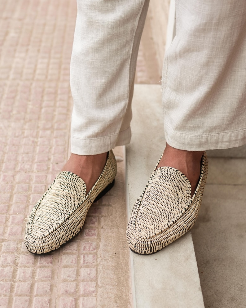 Pode incluir: Um par de mocassins de palha tran&ccedil;ada com acabamento preto. Os mocassins s&atilde;o usados por uma pessoa que usa cal&ccedil;as brancas.