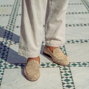 Pode incluir: Um par de mocassins de palha tran&ccedil;ada de cor castanho claro. Os mocassins s&atilde;o usados por uma pessoa em p&eacute; num ch&atilde;o de ladrilhos com um padr&atilde;o de losangos preto e branco.