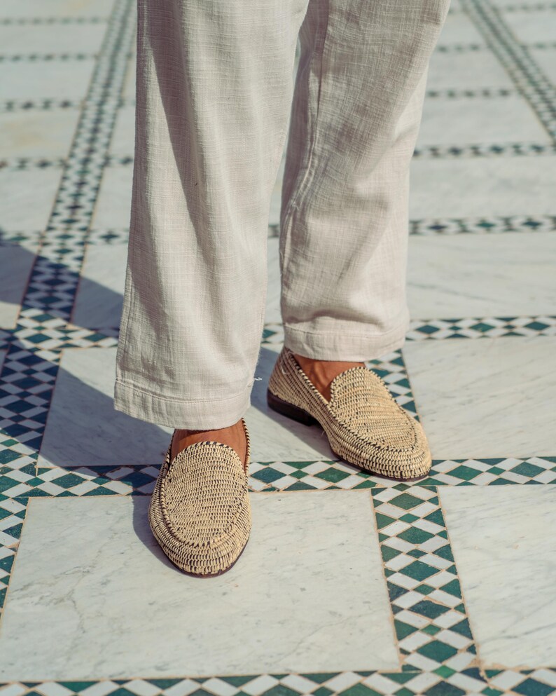 Pode incluir: Um par de mocassins de palha tran&ccedil;ada de cor castanho claro. Os mocassins s&atilde;o usados por uma pessoa em p&eacute; num ch&atilde;o de ladrilhos com um padr&atilde;o de losangos preto e branco.