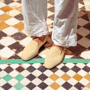 May include: A pair of woven raffia loafers in a light beige color. The shoes are worn with light-colored pants. The shoes have a classic slip-on design and are suitable for casual wear. The shoes are on a checkered tile floor.