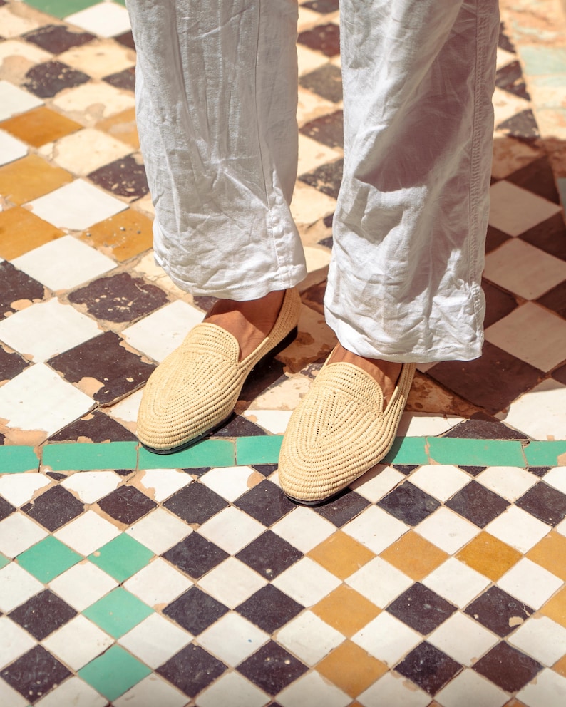 May include: A pair of woven raffia loafers in a light beige color. The shoes are worn with light-colored pants. The shoes have a classic slip-on design and are suitable for casual wear. The shoes are on a checkered tile floor.